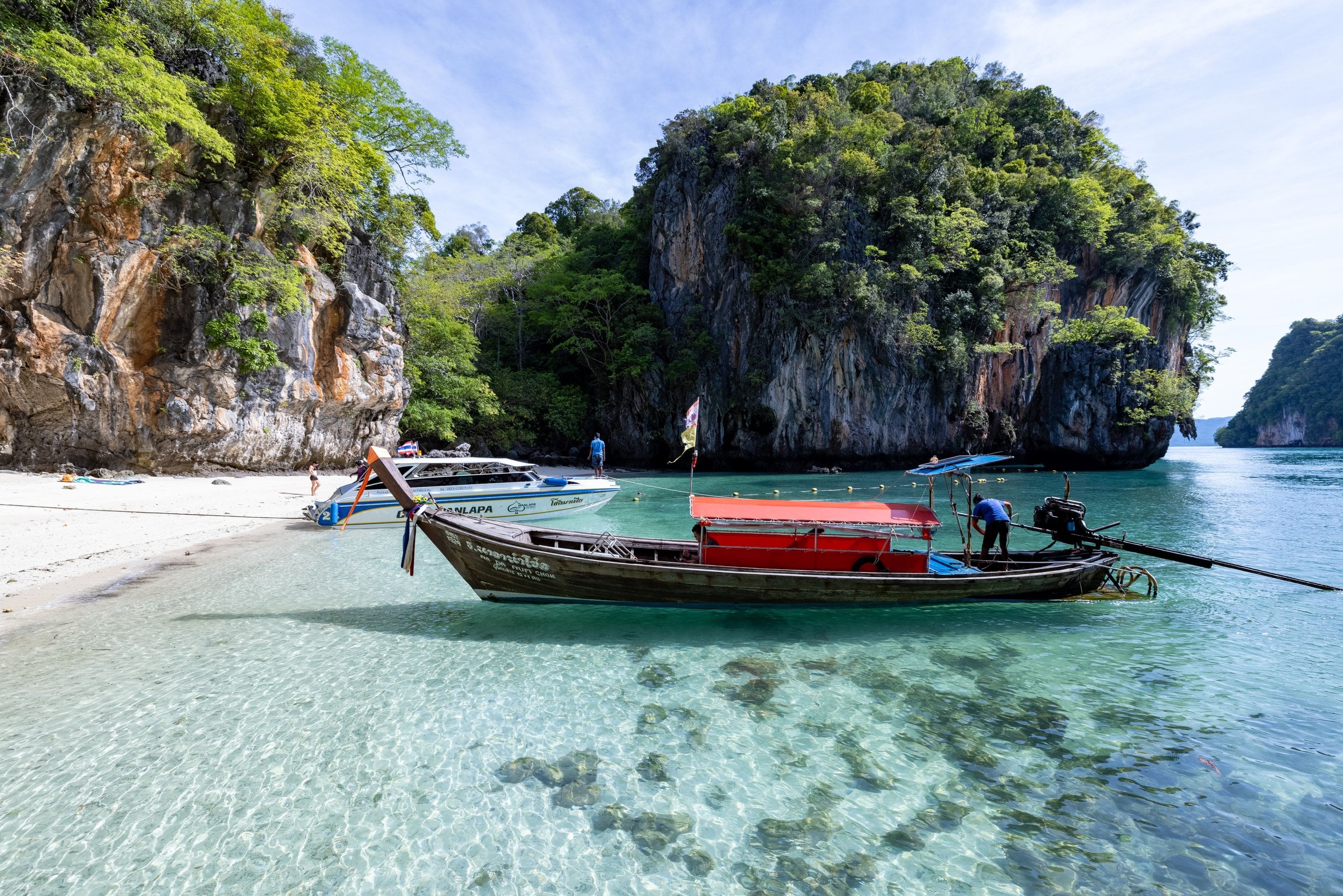 A longtail boat in clear turquoise shallows beside limestone cliffs and a white-sand beach in southern Thailand