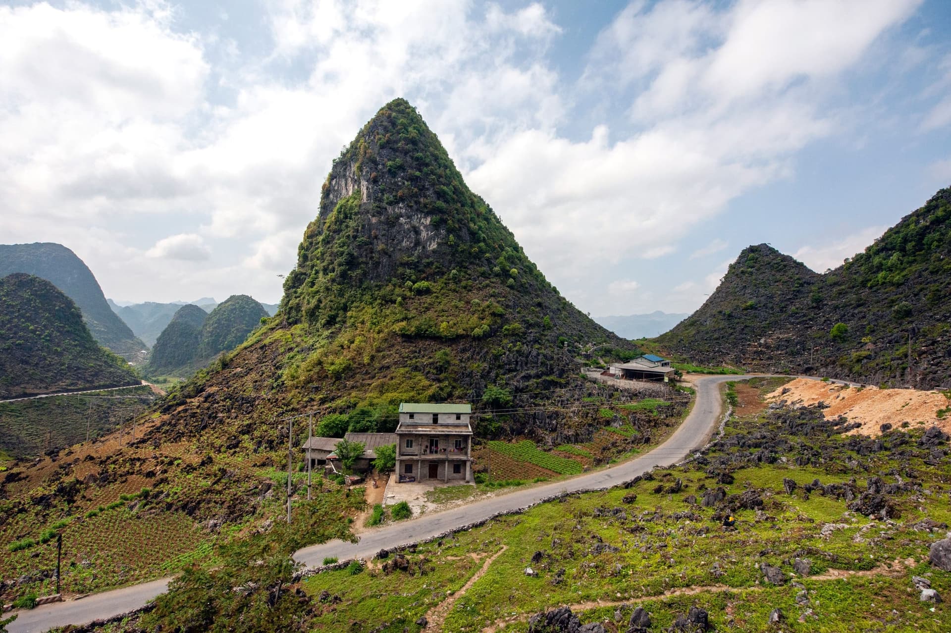 A paved mountain road curving past a farmhouse below a limestone peak in Ha Giang, northern Vietnam. Long mountain legs like this one are why the Hanoi to Sapa transfer is scheduled with two stops.