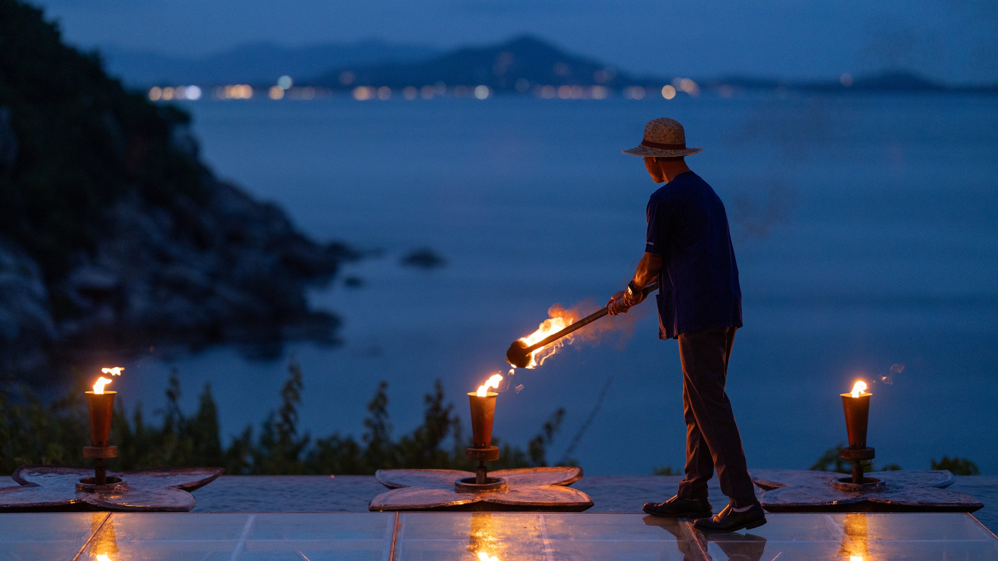 Seorang tuan rumah Banyan Tree Samui menyalakan deretan obor di tepi air saat senja, Koh Samui, Thailand
