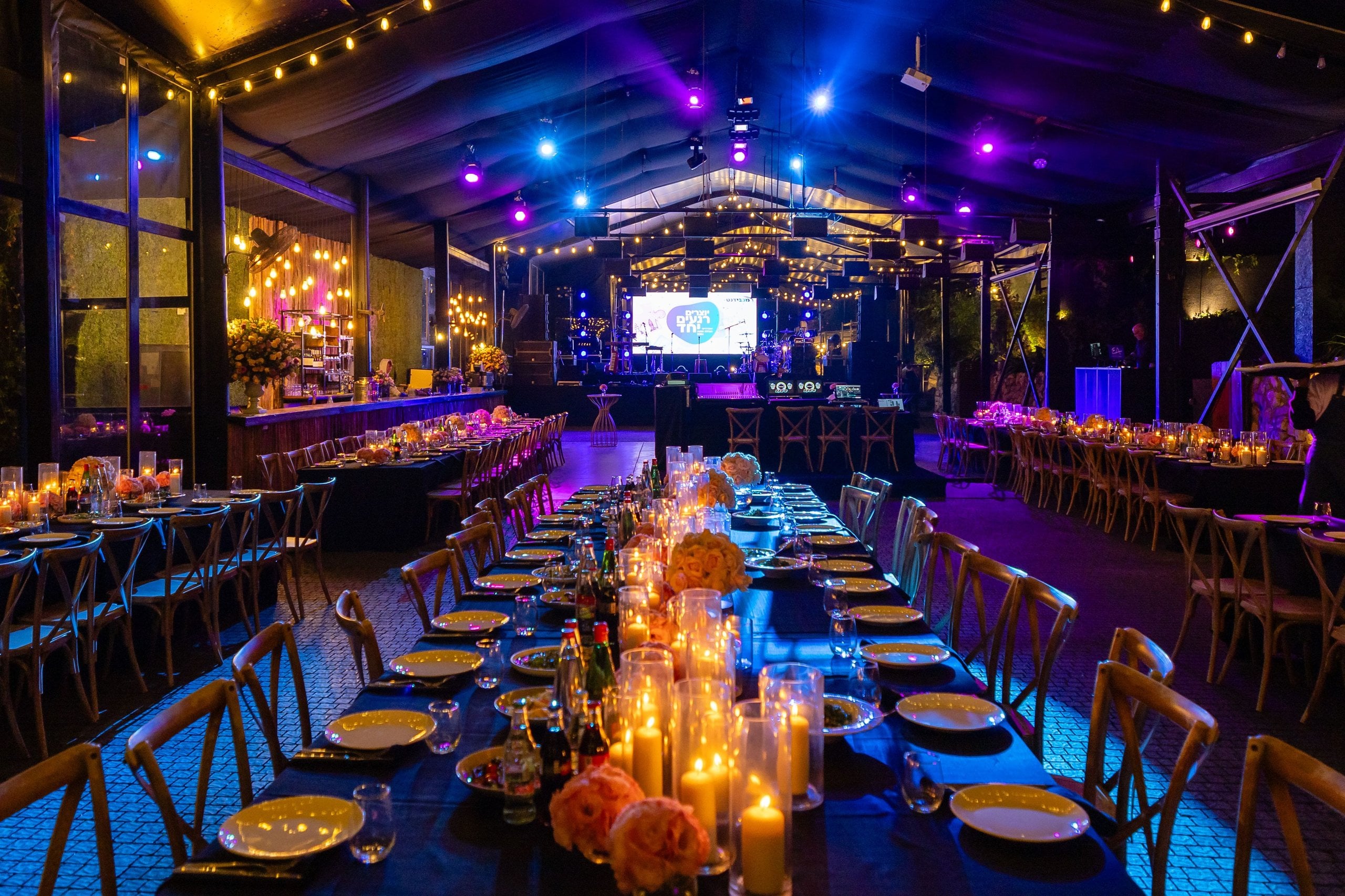 Long candlelit banquet tables under coloured stage lights at an evening event