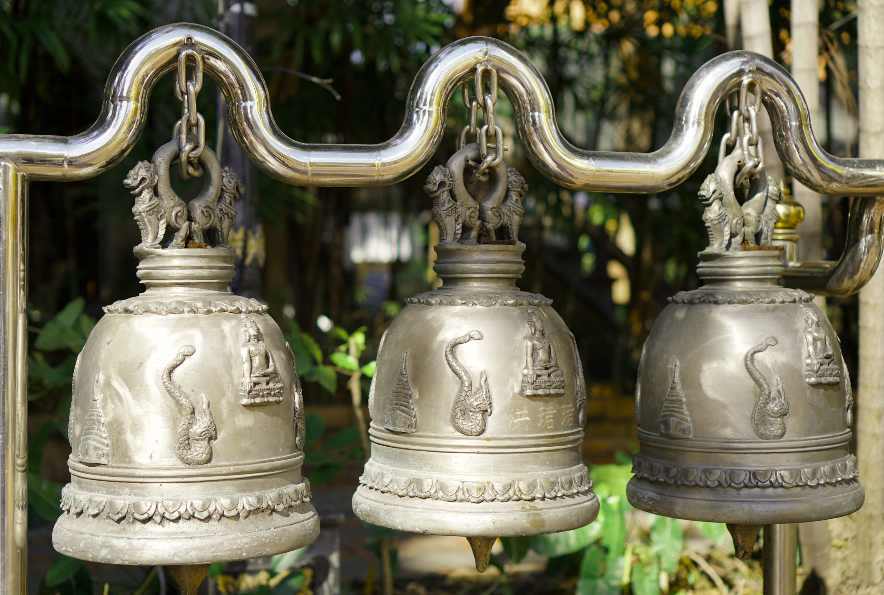 Three cast silver temple bells hanging in a row at a Chiang Mai temple, naga and Buddha figures worked into the metal