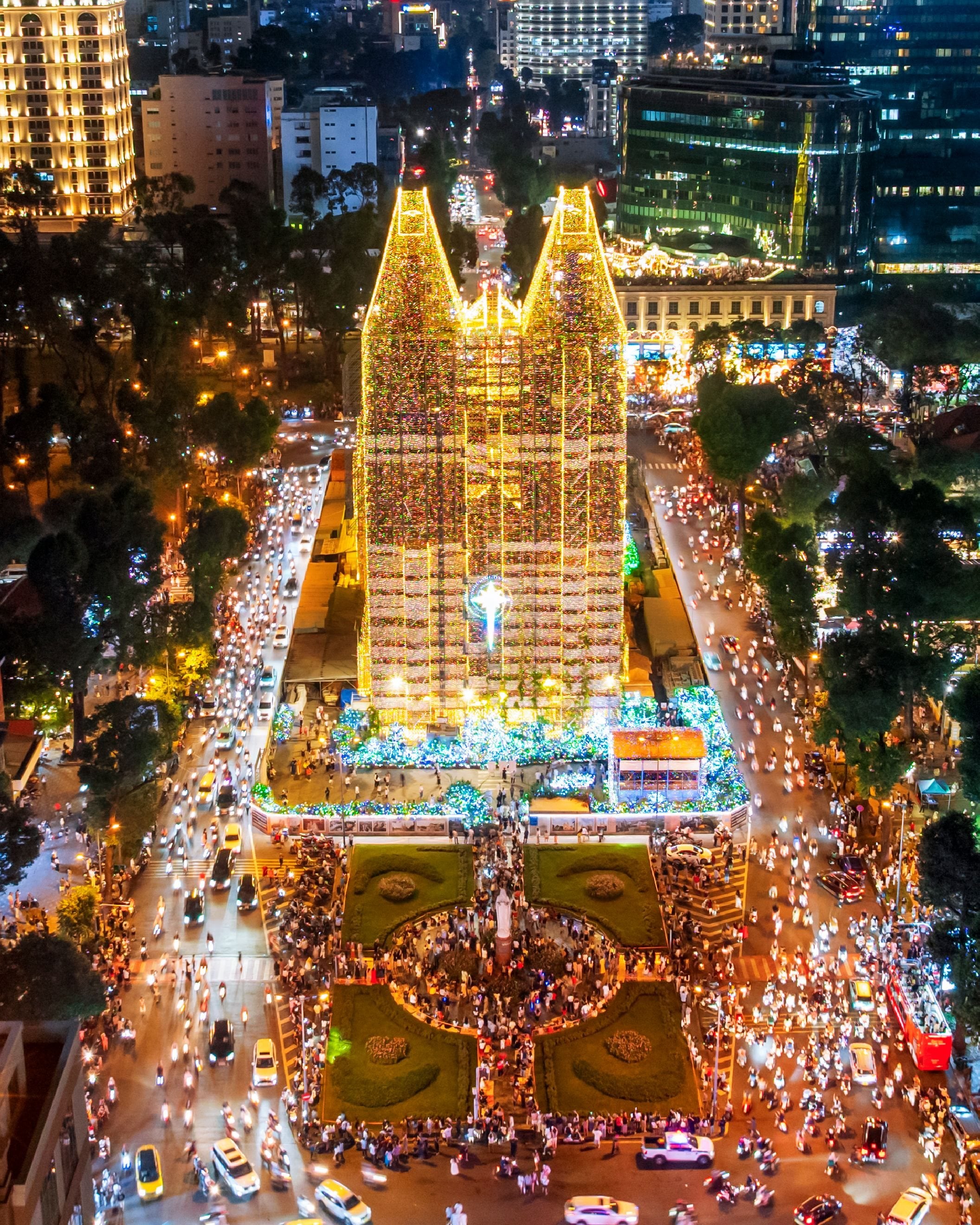 Nhà thờ Đức Bà ở TP. Hồ Chí Minh nhìn từ trên cao vào ban đêm, hai tháp chuông phủ đèn Giáng sinh, quảng trường đông người và xe cộ trên các con phố xung quanh.