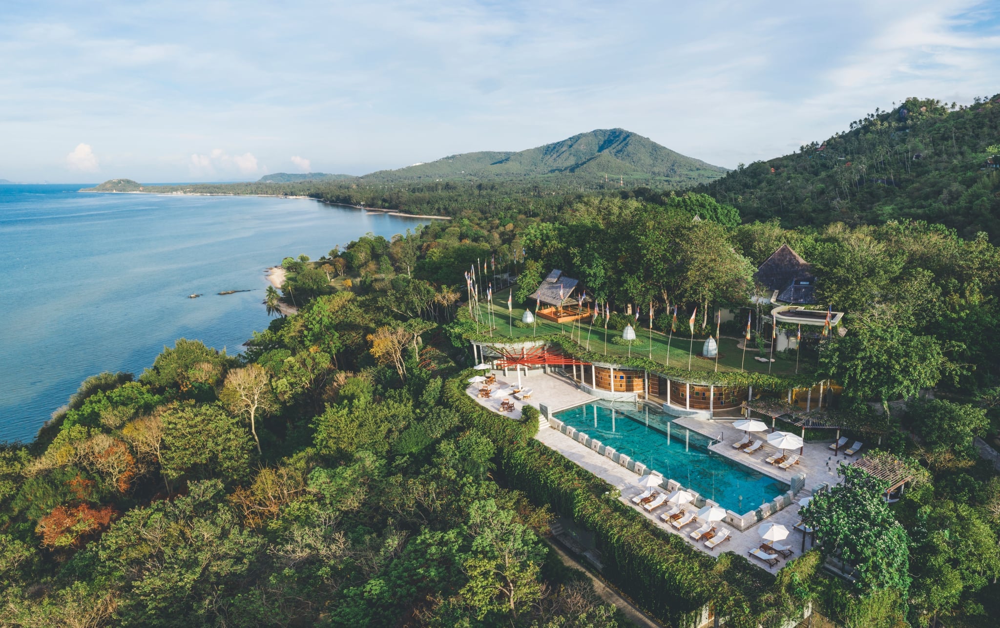An aerial view of Kamalaya's lap pool and lawn on a forested headland, with the Koh Samui coastline and hills behind