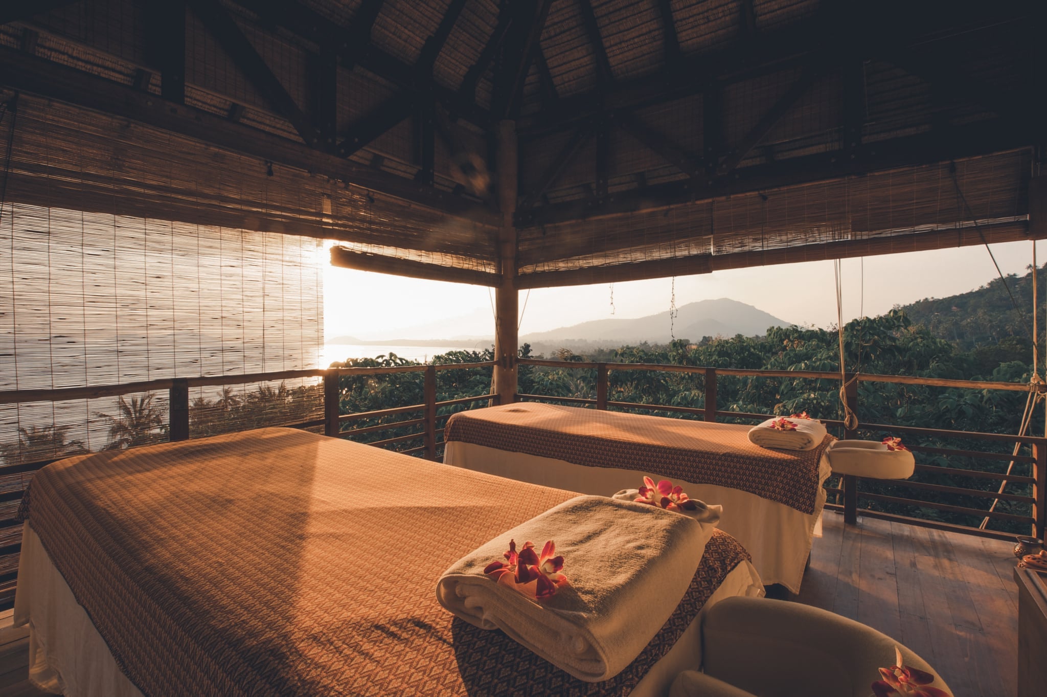 Two treatment beds laid with towels and flowers in an open-sided sala, with forested hills and the sea behind