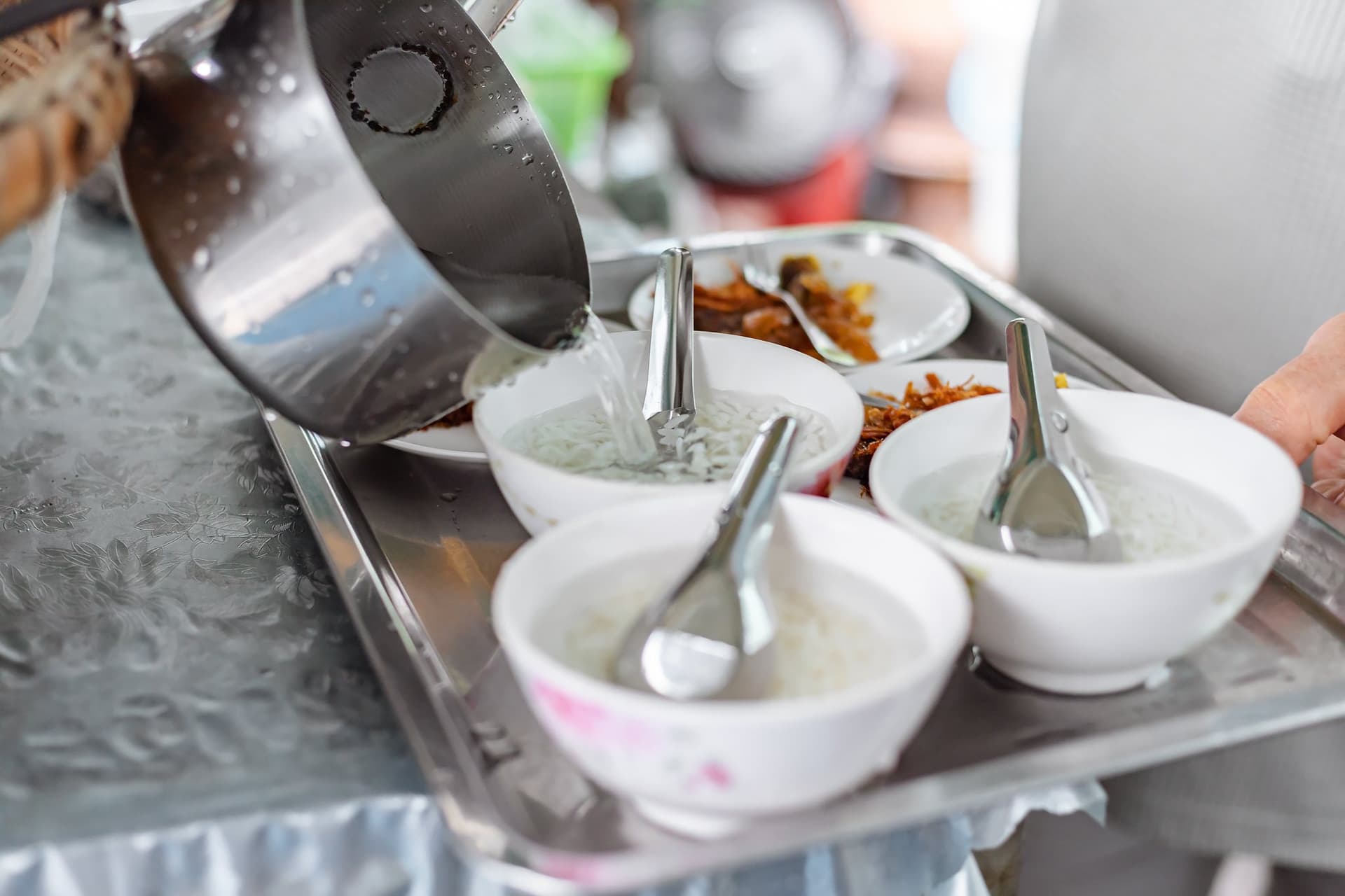 Iced jasmine water being poured from a metal jug into small white bowls of rice, with side dishes on a tray behind.