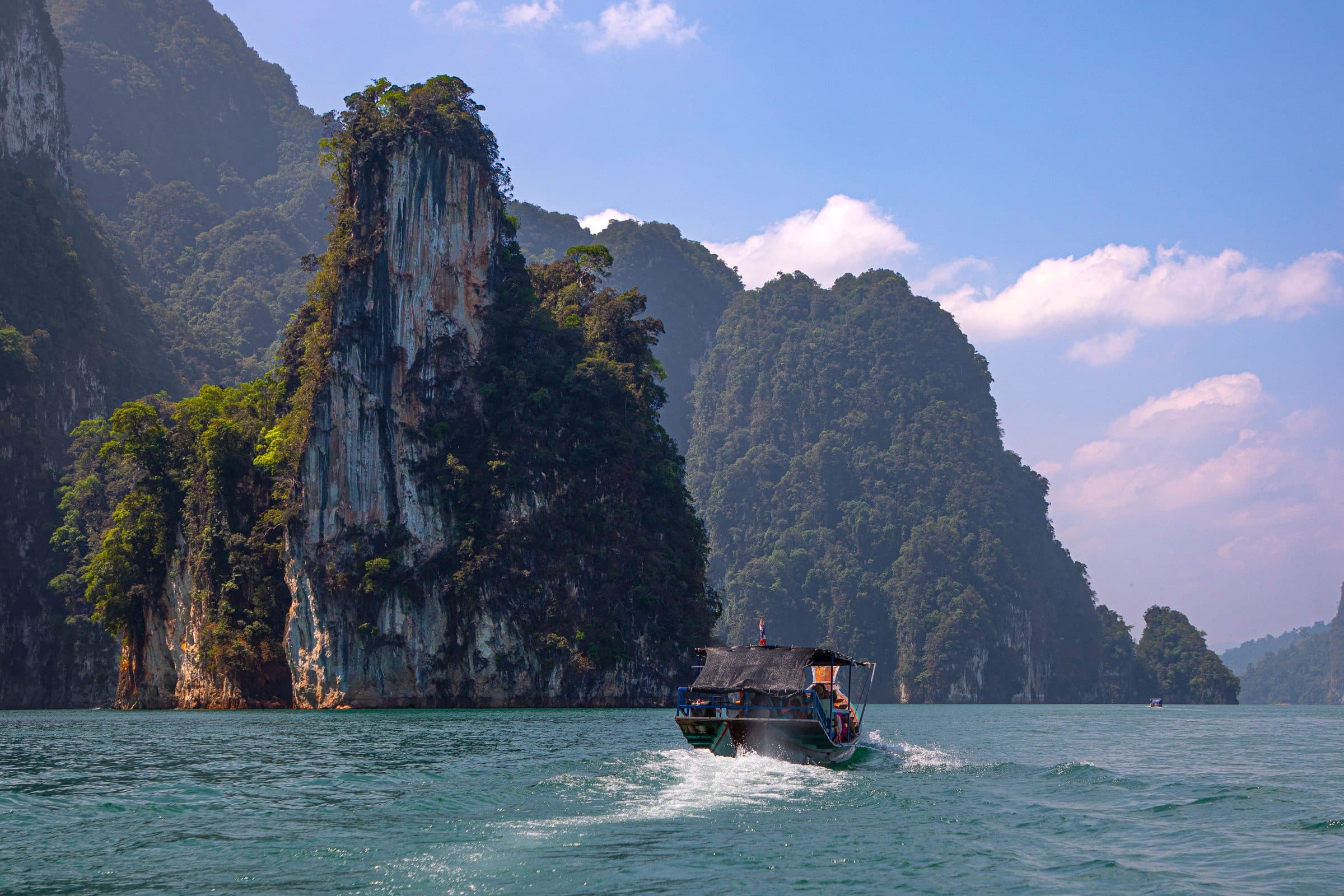 A longtail boat crossing Cheow Lan Lake beneath the limestone cliffs of Khao Sok, Thailand