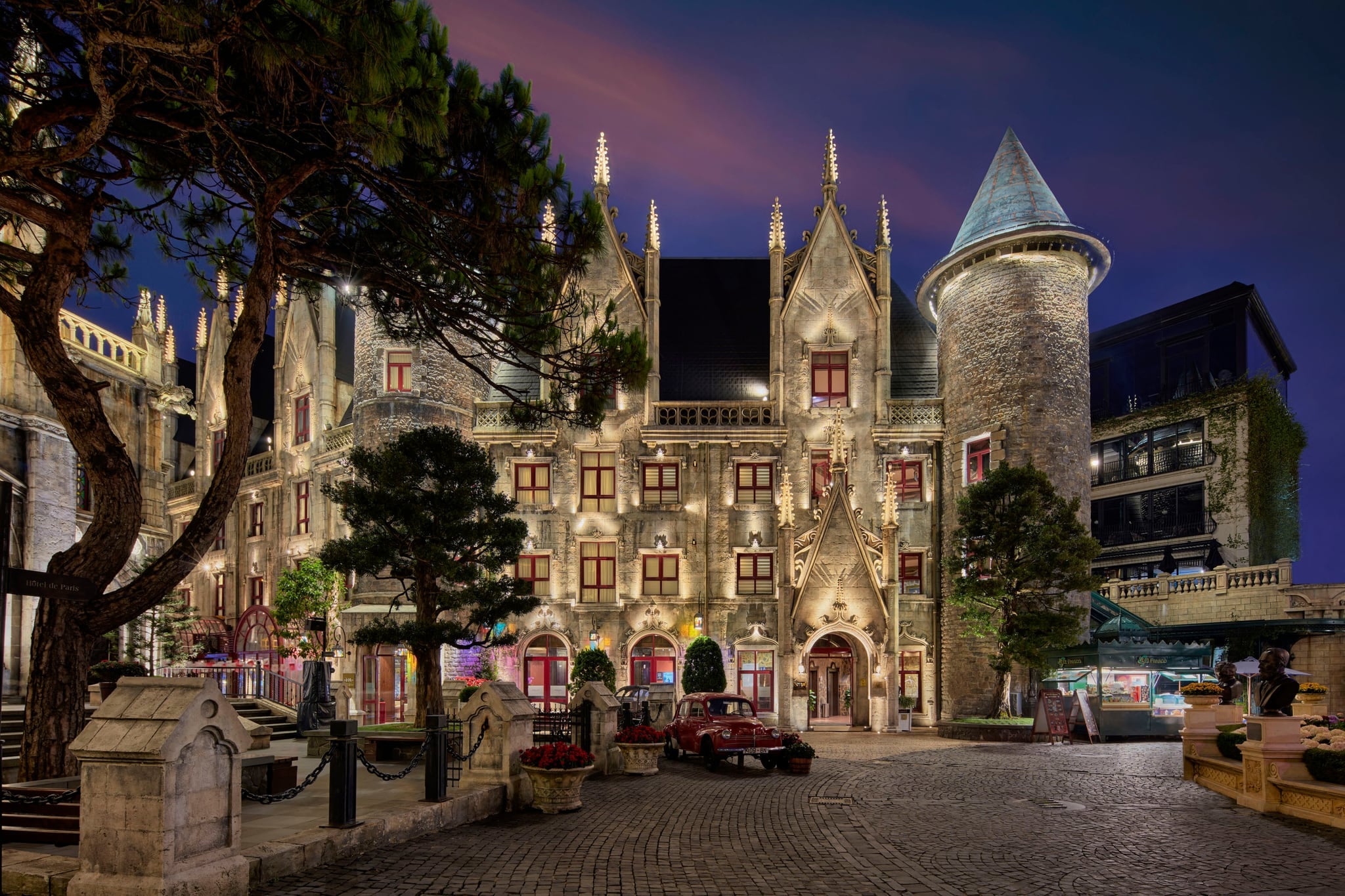 Stone buildings of the French Village at Ba Na Hills lit up at dusk, with turrets, red-framed windows and pine trees
