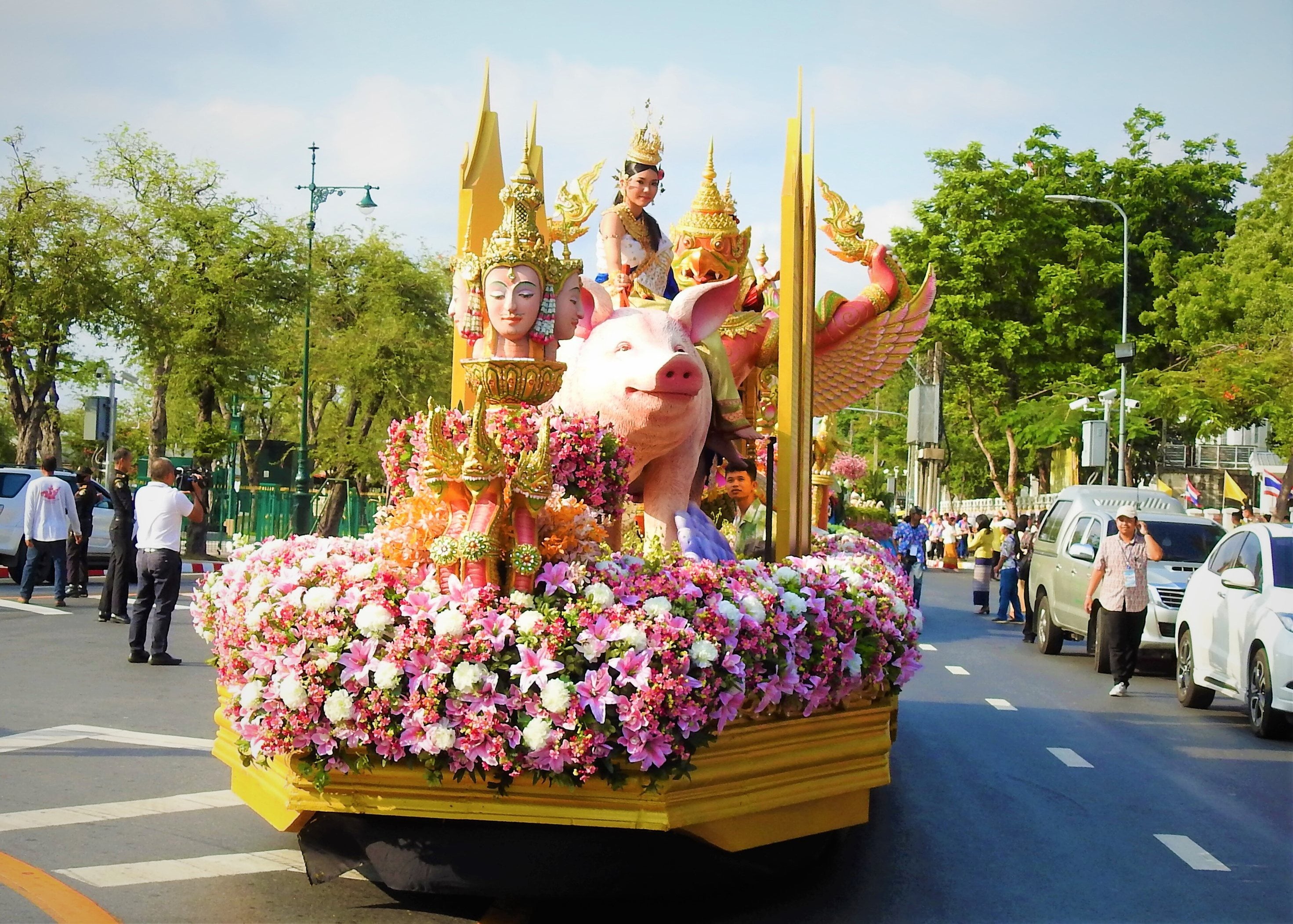 A parade float covered in pink and white flowers carrying a crowned woman riding a large pig, with gilded figures beside her, on a city road.