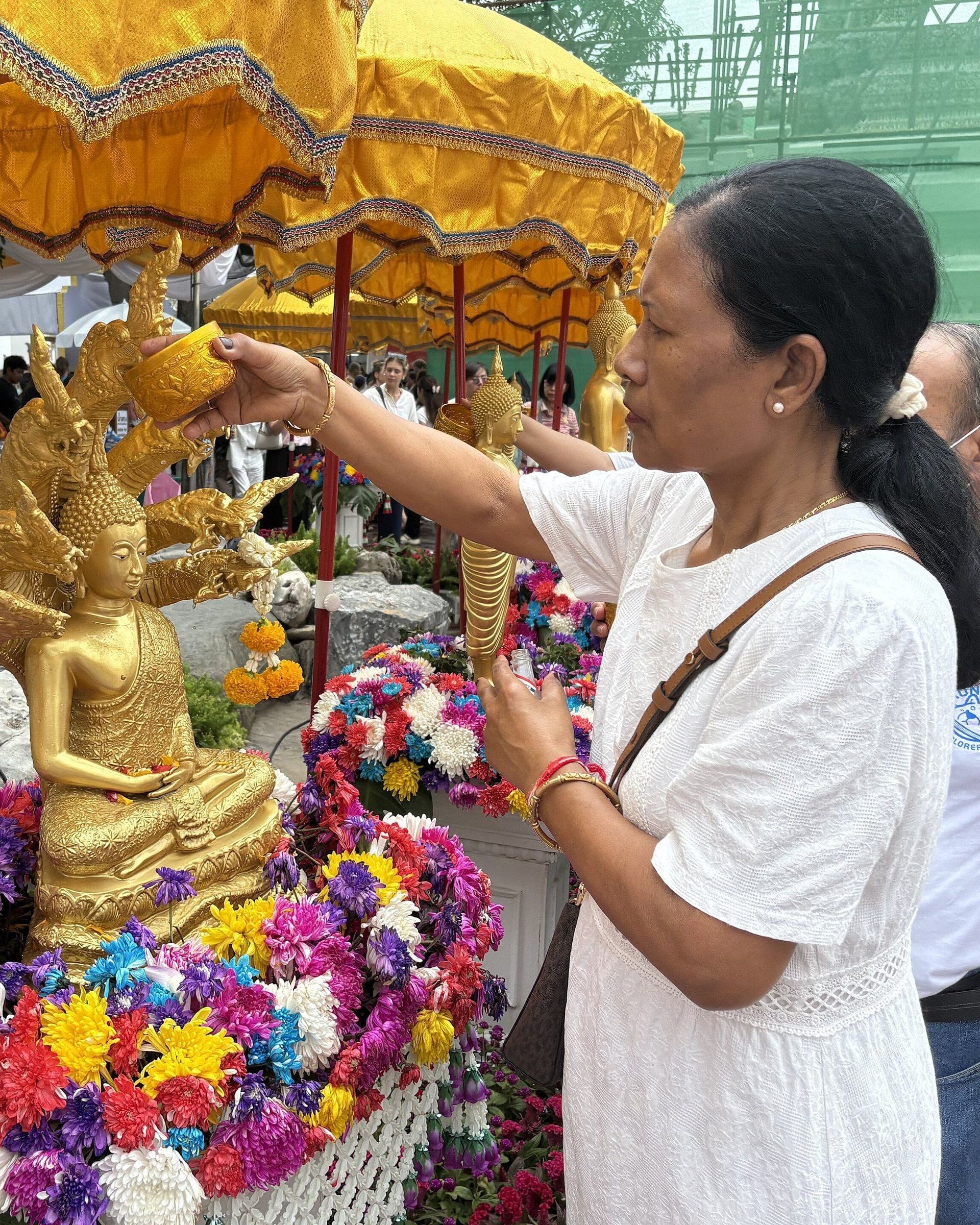 A woman in white pouring water from a small bowl over a gilded Buddha image sheltered by a naga and ringed with bright flowers, under yellow umbrellas.