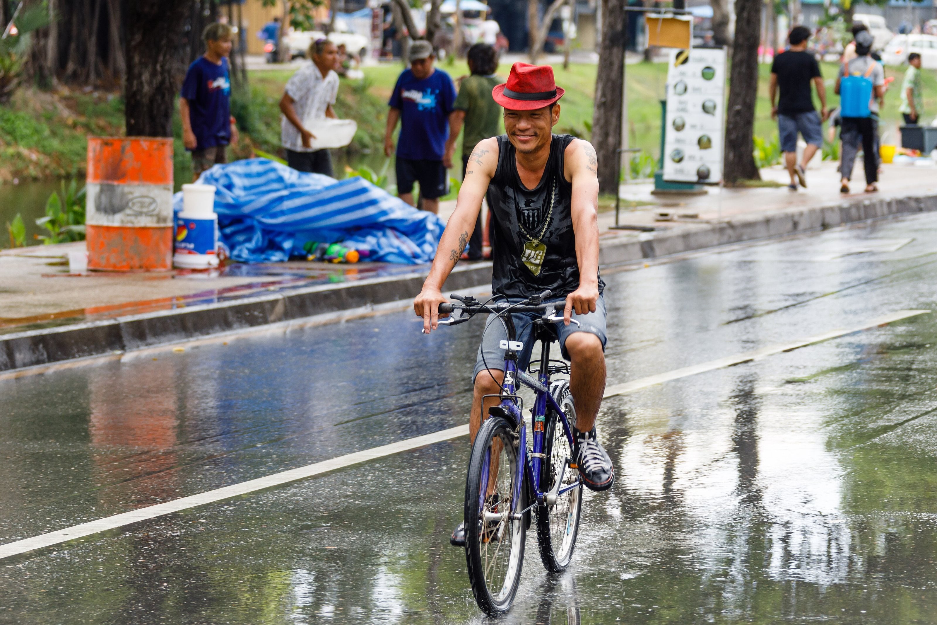 A smiling man in a red hat and a black vest cycling along a soaked road beside the Chiang Mai moat, with people carrying water along the verge.