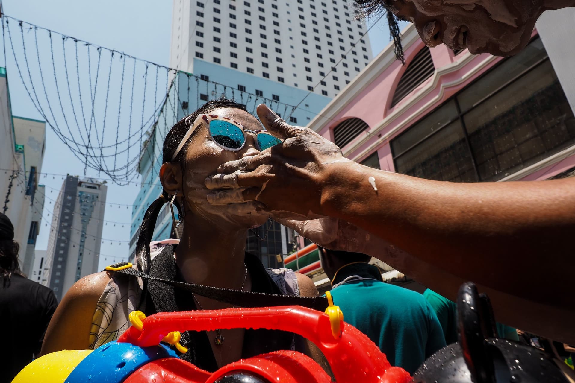Hands smoothing white paste onto the face of a woman in blue sunglasses, with a plastic water gun in front of her.