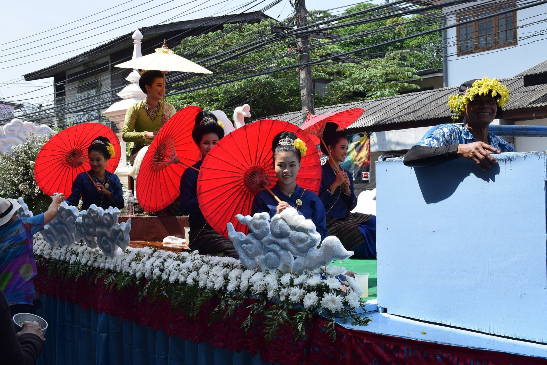 Young women in dark blue dress holding red paper parasols on a float decked with white flowers, with a white swan figure behind them.