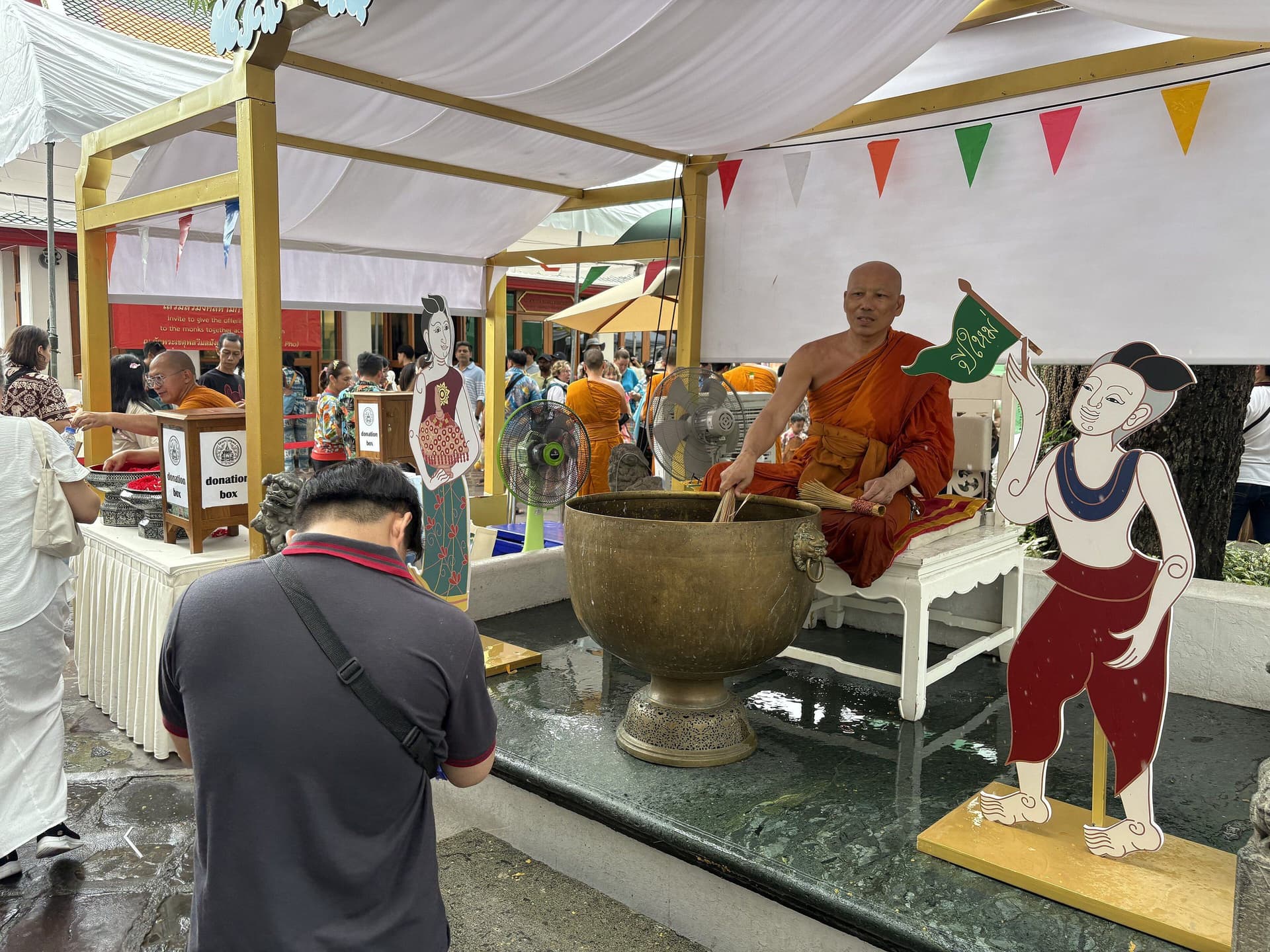 A monk in an orange robe seated beside a large brass bowl of water under a white canopy, with painted cut-out figures beside him and a visitor waiting in front.