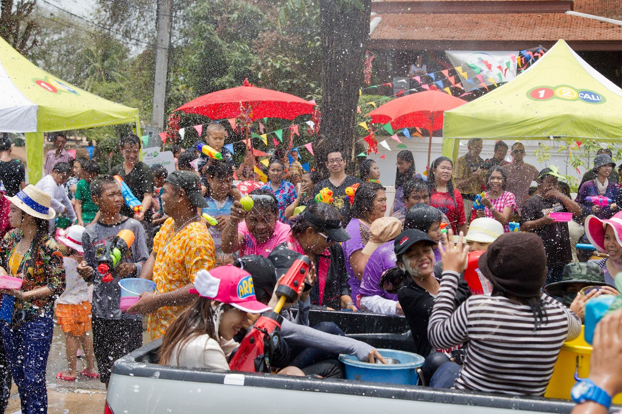 People with water guns and bowls crowding around and into the back of a pickup truck as water flies, under market umbrellas and bunting.