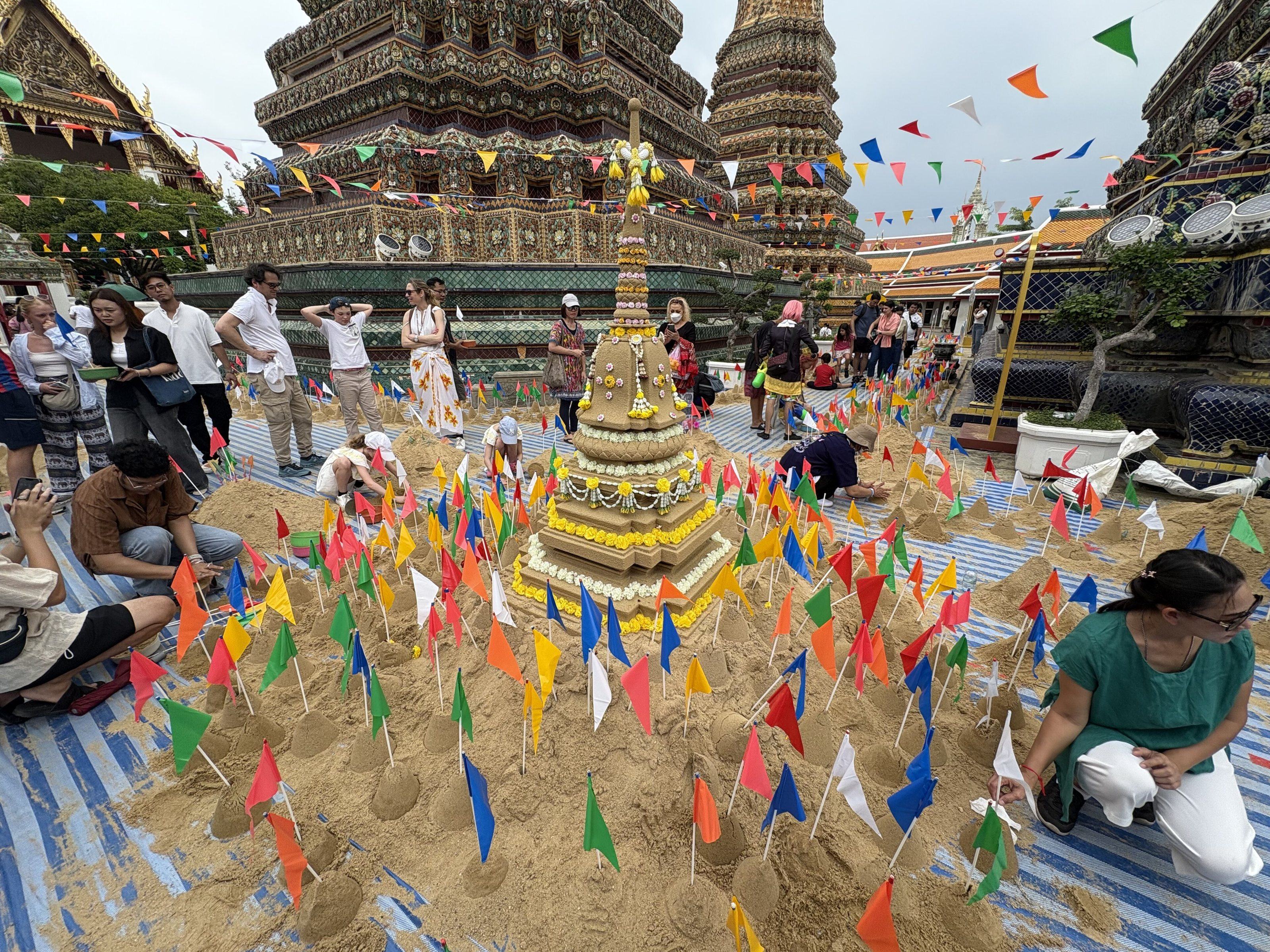A tiered sand chedi wrapped in yellow and white garlands, surrounded by hundreds of small coloured paper flags planted in sand, with the tiled chedis of Wat Pho behind.