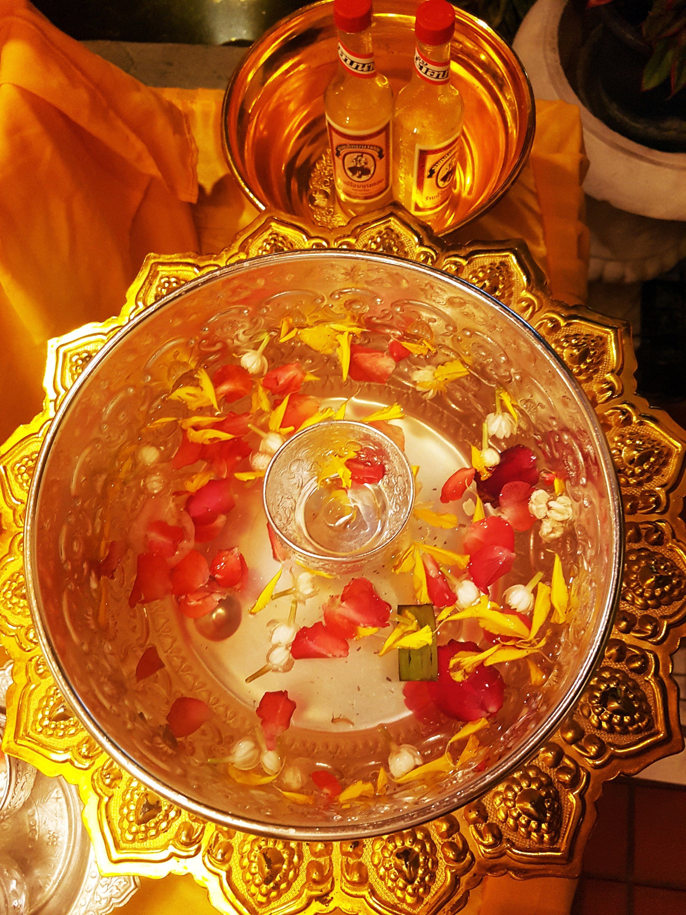 A silver bowl of clear scented water scattered with red and yellow petals and jasmine buds, set on an ornate gold stand.