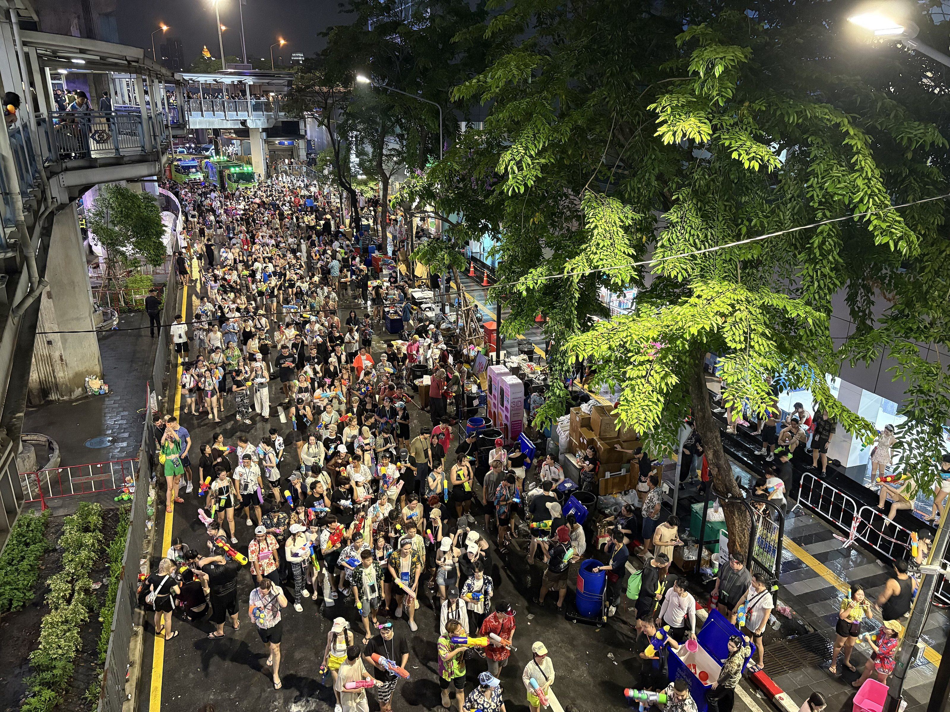 Silom Road at night packed from side to side with people in a water fight, seen from above under street lights and trees.