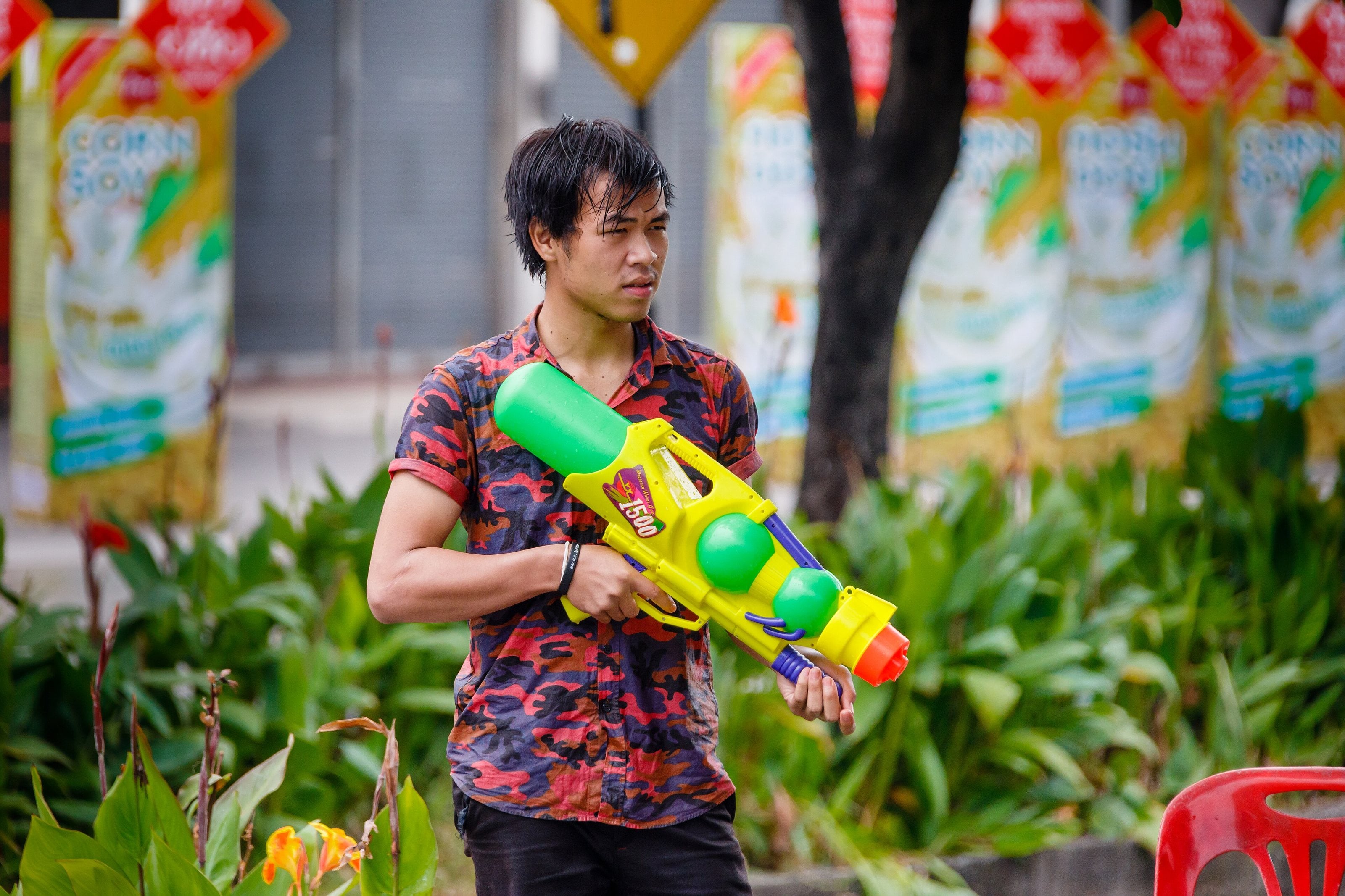 A young man in a red patterned shirt holding a large green and yellow water gun, with festival banners hanging behind him.
