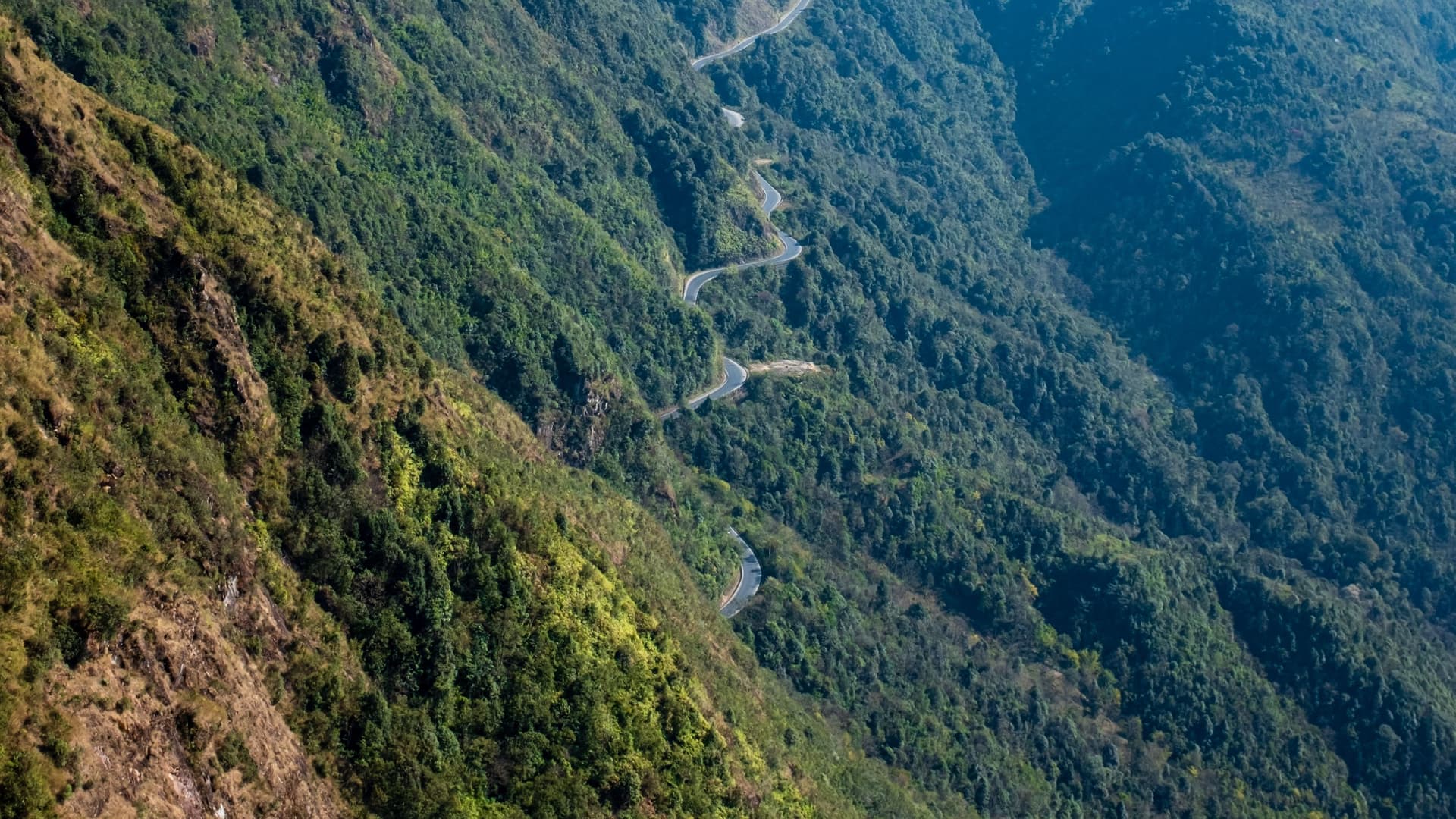 An empty mountain road switchbacking down a forested valley. Ground operations run on roads like this, which is the reality a polished itinerary cannot stand in for.