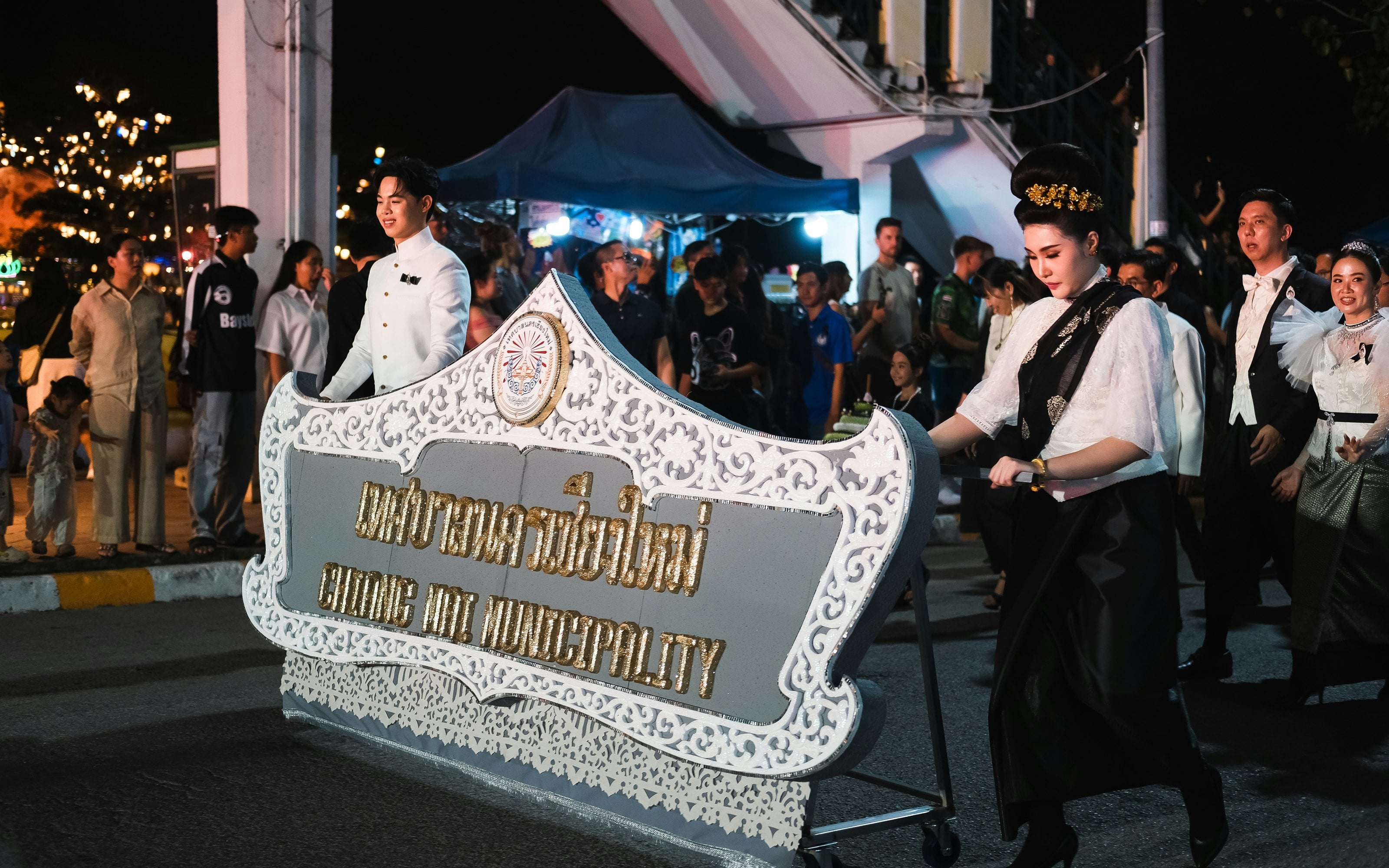 Trên đường phố về đêm, những người diễu hành trong trang phục đen trắng đẩy một tấm biển trắng chạm hoa văn mang tên chính quyền thành phố Chiang Mai bằng tiếng Thái và tiếng Anh, khán giả đứng xem bên lề đường.