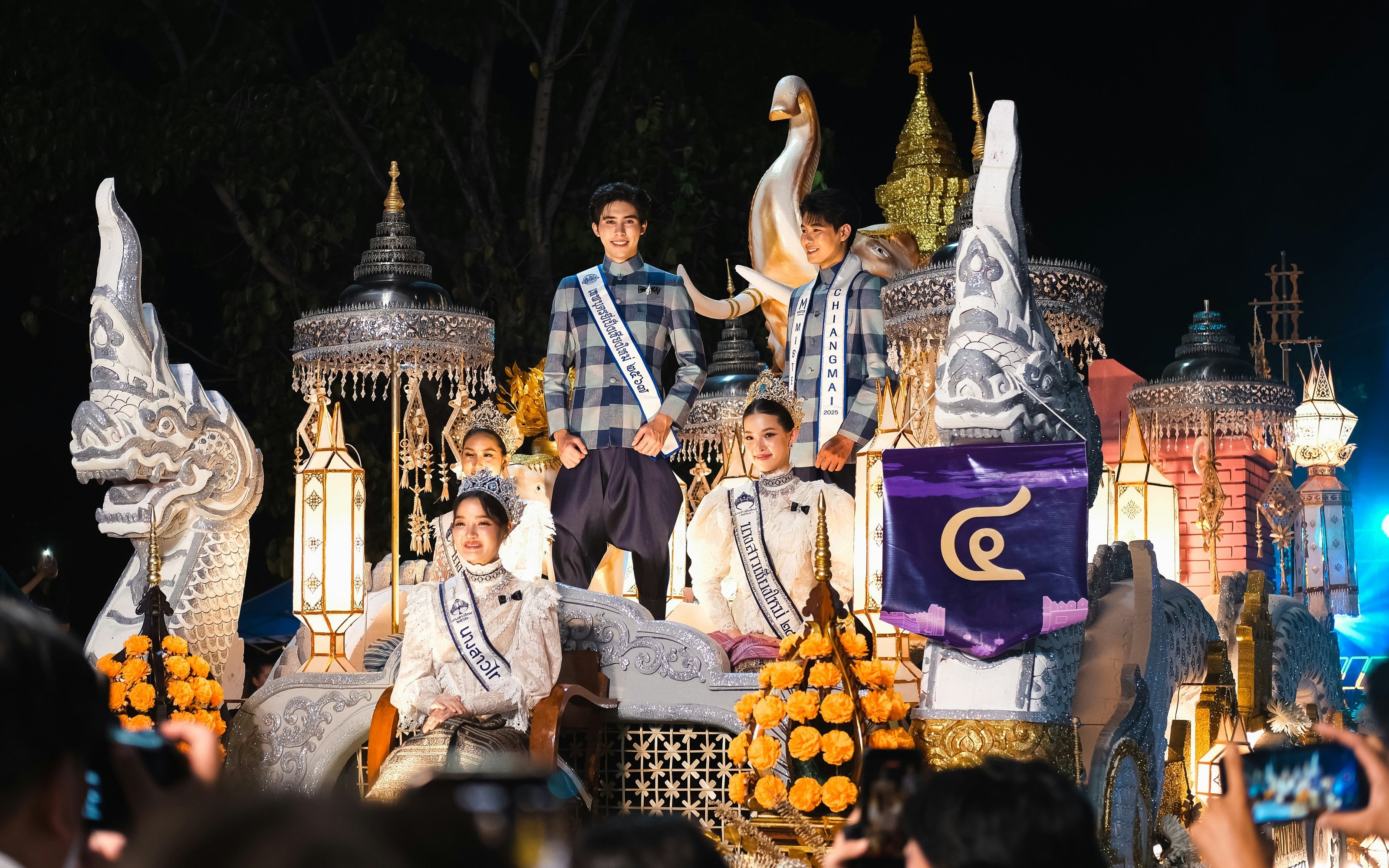A lit parade float at night with white naga heads, silver parasols and marigolds, carrying young men and women in sashes, the women also wearing crowns, as people in the crowd hold up their phones.