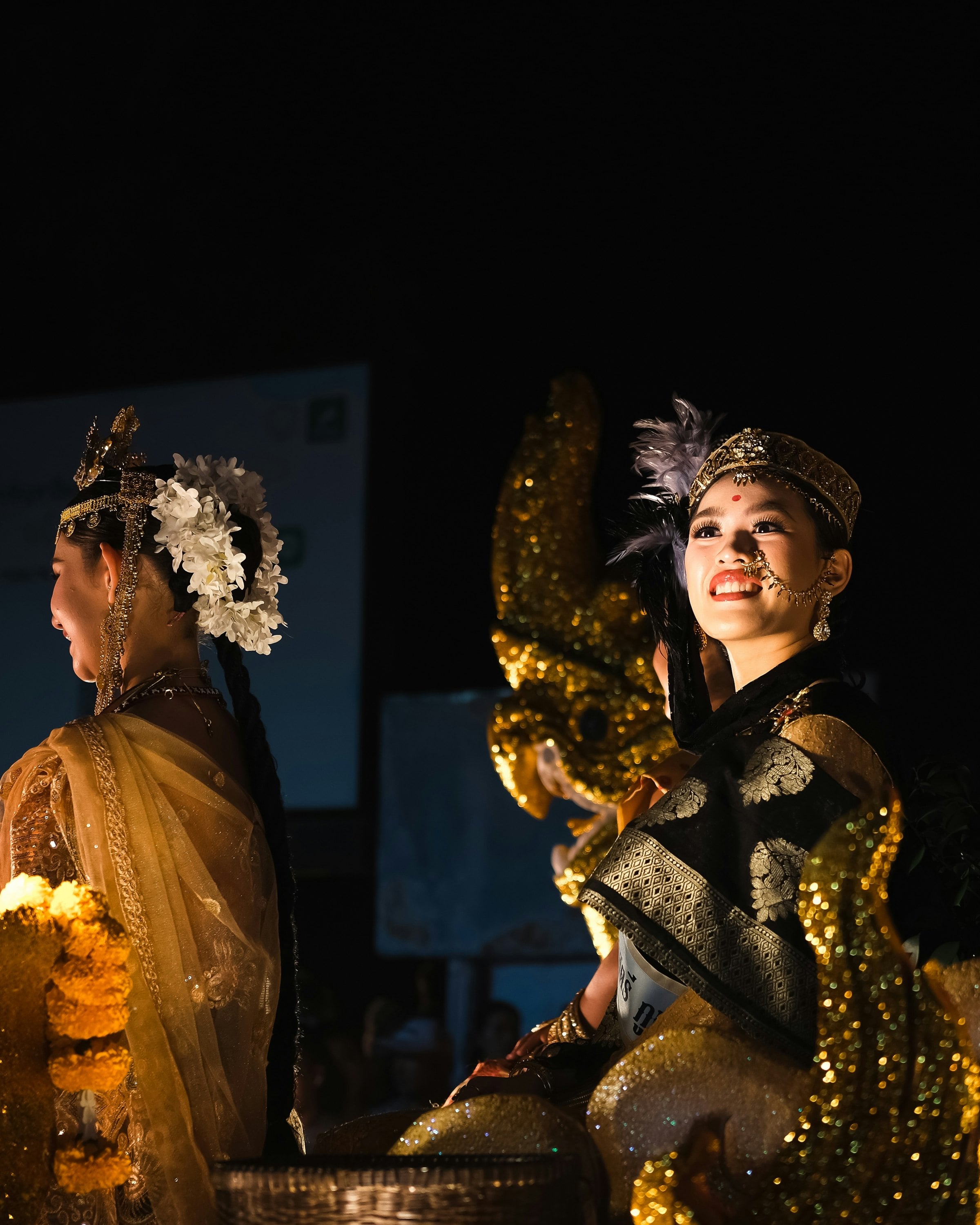 Hai cô gái trong trang phục cầu kỳ trên một xe hoa về đêm: một người nghiêng mặt, cài hoa trắng trên tóc; người kia mặc đồ đen ánh vàng, mỉm cười ngước nhìn lên bên cạnh một đầu rắn thần naga vàng óng.