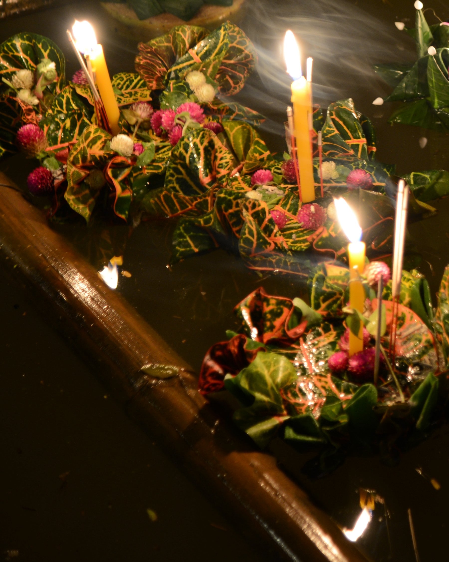 Những chiếc krathong gấp từ lá cô tòng, cắm nến và nhang đang cháy, trôi trên mặt sông tối bên một cây sào tre.